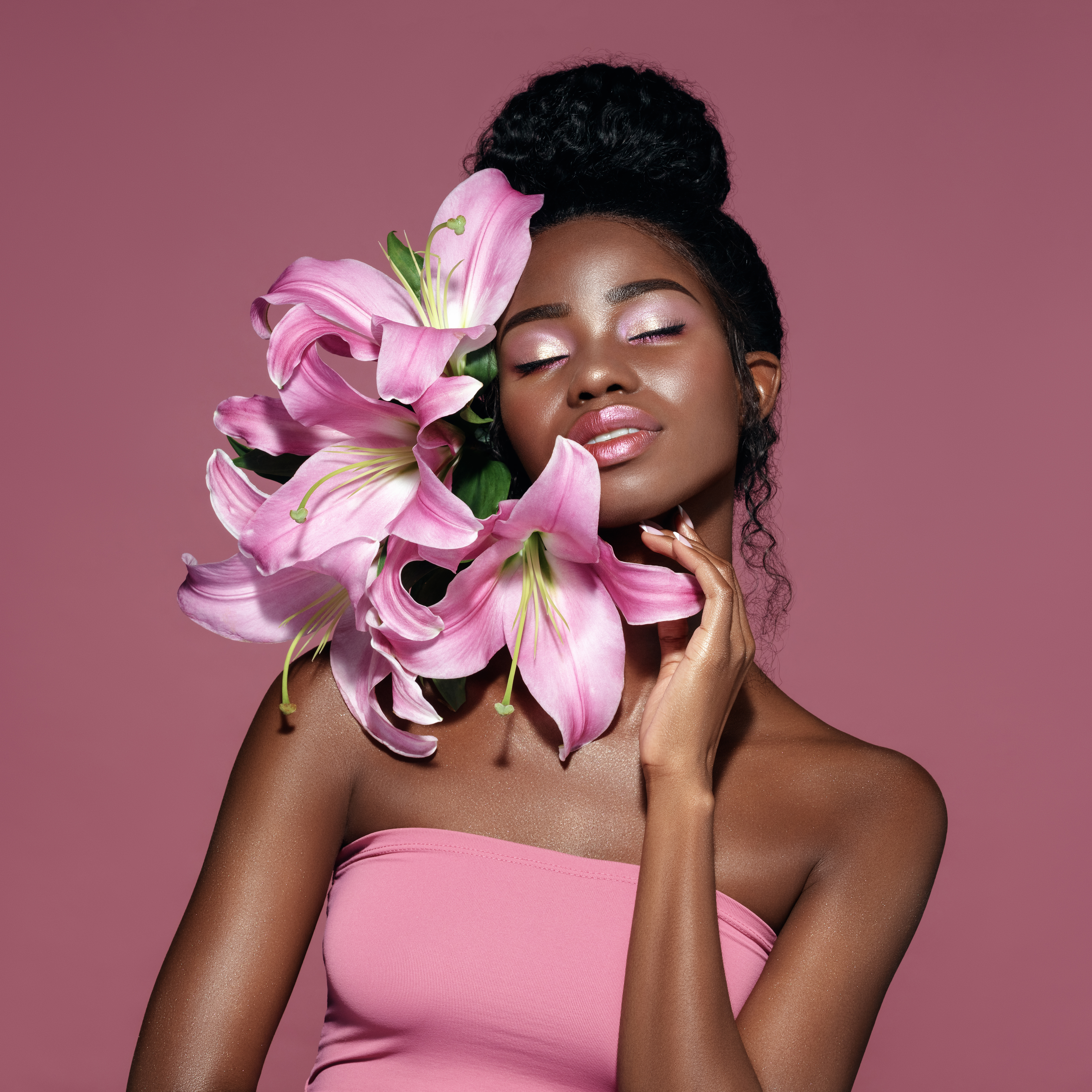 Fashion beauty portrait of young African American model with pink art make up posing with lily flowers against pink background.